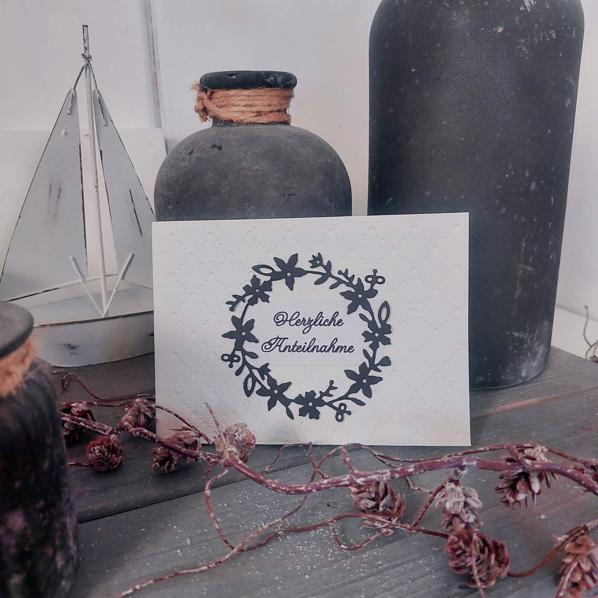 Die Stanzschablone Blumenkranz von Stanzenshop.de dekoriert mit "Herzliche Anteilnahme" liegt zwischen dunklen Vasen und Zweigen auf rustikalem Holztisch - ideal zum Basteln und Gestalten von Trauerkarten.