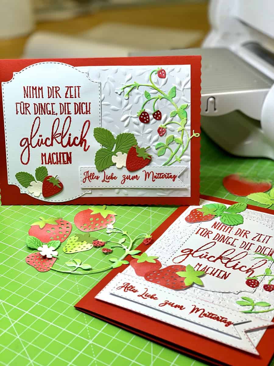 Zwei handgefertigte Grußkarten mit Erdbeeren und Blättern, hergestellt mit der "Stanzschablone Erdbeeren" von Stanzenshop.de, mit deutschem Text auf einer grünen Schneidematte mit Bastelwerkzeug im Hintergrund.