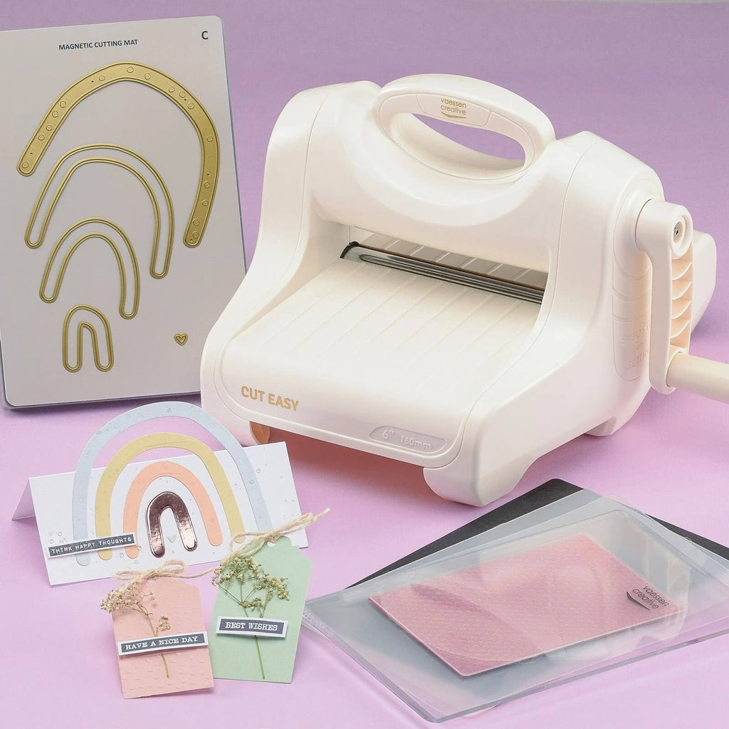 Eine Vaessen Creative • Cut Easy Stanz- und Prägemaschine A5 von Sizzix ist zusammen mit Zubehör wie Stanzformen, farbigem Papier und verschiedenen Bastelmaterialien auf einer hellvioletten Oberfläche angeordnet.