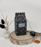 Ein schwarzer Geschenkkarton mit weißem Baummotiv, hergestellt mit der Stanzschablone Schachtel mit Öffnung von Stanzenshop.de, ruht auf einem Holzpodest, umgeben von getrockneten Pflanzen in einer Vase, einem Zweig und einem künstlichen Herbstblatt.