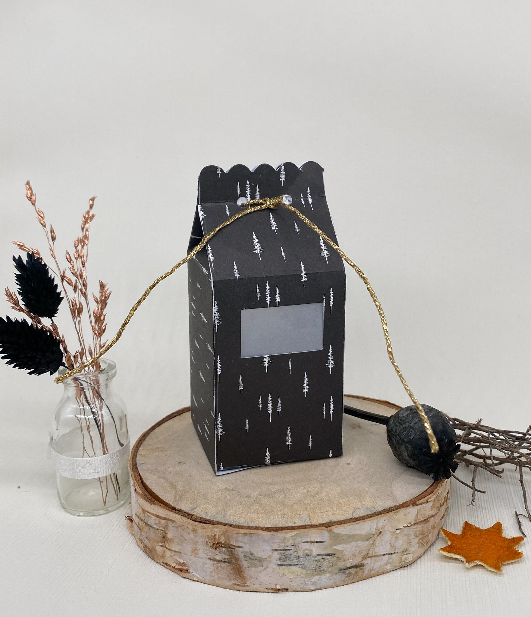 Ein schwarzer Geschenkkarton mit weißem Baummotiv, hergestellt mit der Stanzschablone Schachtel mit Öffnung von Stanzenshop.de, ruht auf einem Holzpodest, umgeben von getrockneten Pflanzen in einer Vase, einem Zweig und einem künstlichen Herbstblatt.