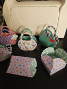 Stanzschablone Geschenkkästchen von Stanzenshop.de: Bunte, kleine Papier-Geschenkkästchen mit verschiedenen Mustern und Formen - ideal zum Basteln oder Scrapbooking - auf dunkler Fläche vor weißem Behälter ausgestellt.