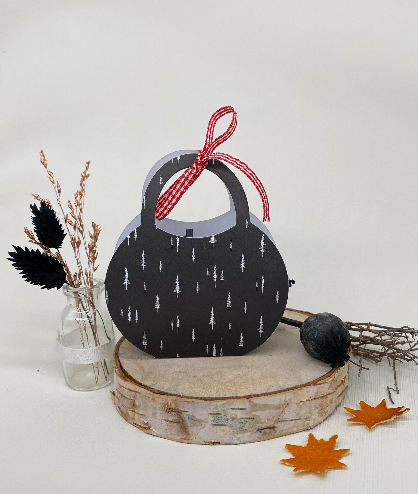 Eine schwarze, runde Papiertüte mit weißen Baummotiven und rotem Band aus der Stanzschablone Bastelset Handtasche von Stanzenshop.de liegt auf einer Holzplatte zwischen Herbstblättern und Trockenblumen - ideal zum Basteln oder Scrapbooking.