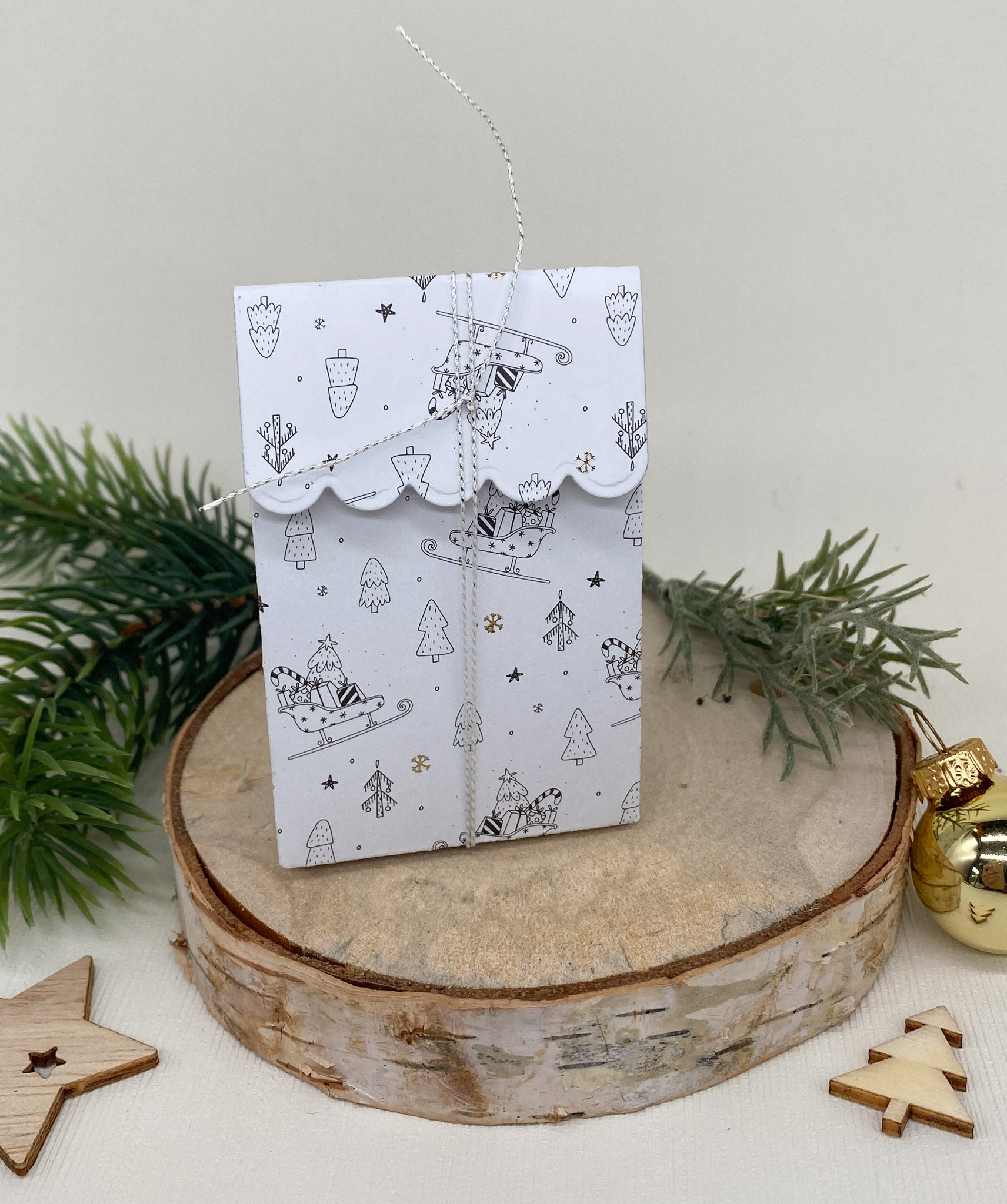 Ein weißer Geschenkumschlag mit winterlichen Motiven und Kordelverschluss ist auf Holz mit Tannenzweigen und Sternen abgebildet - die perfekte Inspiration für Gastgeschenke oder Bastelboxen mit der "Stanzschablone Schachtel mit Verzierungen" von Stanzenshop.de.