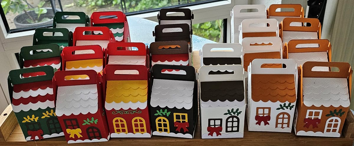 Ein Set bunter, hausförmiger Geschenkboxen mit dem Stanzschablone Bastelset für eine Schachtel mit winterlichen Motiven von Stanzenshop.de, mit einzigartigen Dach- und Türmotiven, arrangiert auf einem Tisch an einem Fenster.