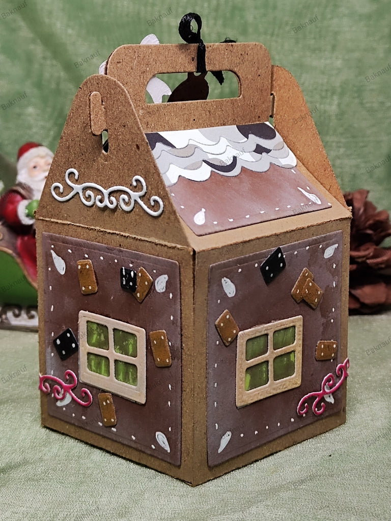 Eine kleine Schachtel mit Lebkuchenmotiv und Griff, die einem Haus ähnelt und mit dekorativen Details wie Fenstern, Zuckerguss und Süßigkeiten verziert ist. Im Hintergrund ist eine Weihnachtsmannfigur verschwommen, die das gemütliche Wintermotiv dieses bezaubernden Stanzschablonen-Bastelsets für eine Schachtel mit winterlichen Motiven von Stanzenshop.de unterstreicht.