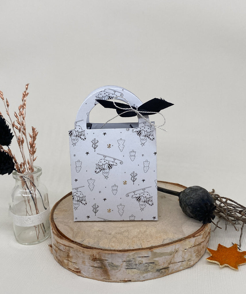 Eine verzierte weiße Geschenktüte mit schwarz-weißem Design liegt auf einer Holzplatte, perfekt für Scrapbooking-Projekte. Daneben steht ein Glasgefäß mit Trockenblumen und einem Trockenblumenarrangement auf der Oberfläche.
