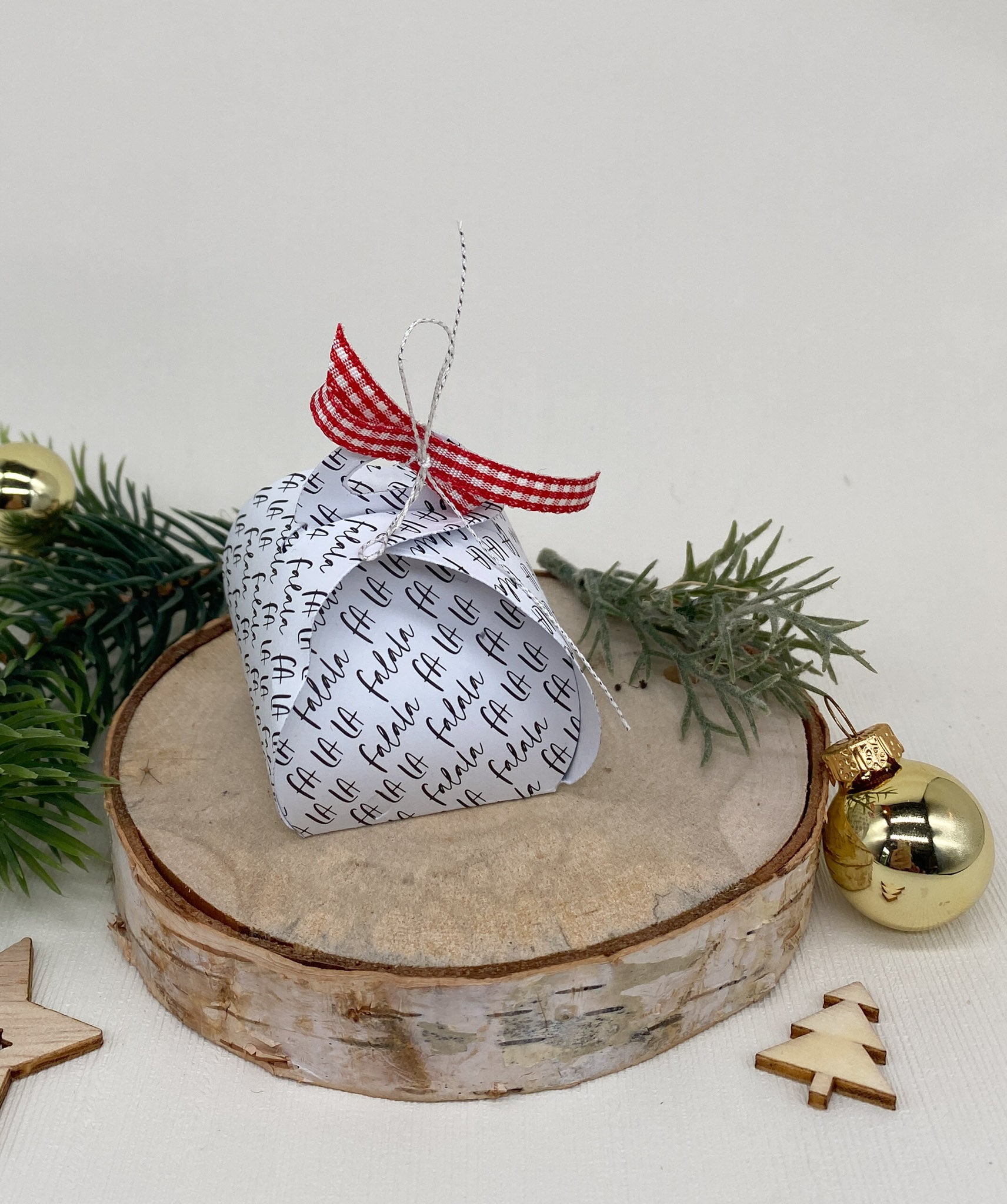 Ein Stanzschablone Schmuckkästchen von Stanzenshop.de mit schwarzem Text und einem rot karierten Band sitzt auf einer Holzplatte, umgeben von Tannenzweigen, einem goldenen Ornament und Holzdekorationen.