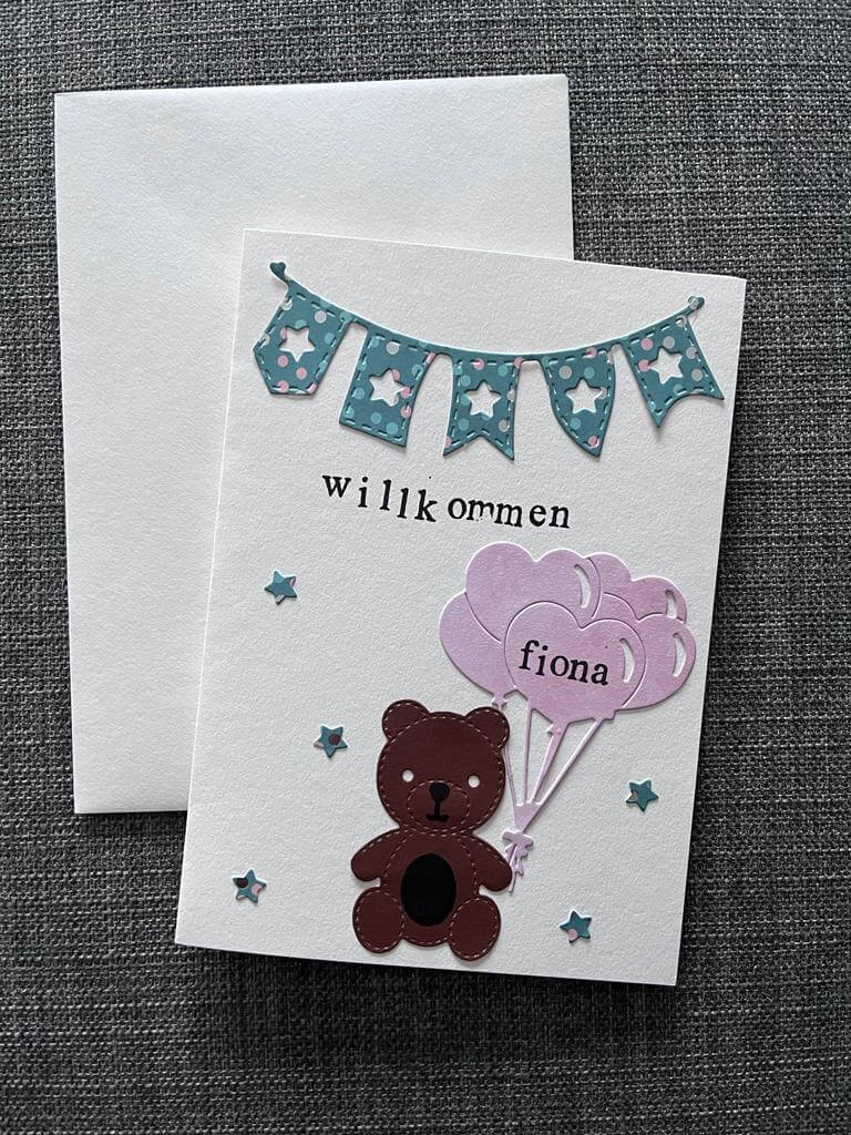 Eine entzückende Geburtstagskarte für Scrapbooking-Fans mit der Stanzschablone Wimpelkette von Stanzenshop.de, einem Bären mit rosa "Fiona"-Luftballons, blauen Wimpeln, "Willkommen"-Schriftzug auf grauem Stoff und einem Umschlag, der dahinter steckt.