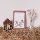 Eine handgefertigte Karte mit der Stanzenshop.de "Stanzschablone Fahrrad mit Blumen", gepaart mit einem Holzhaus und einer Trockenblumenvase auf Stroh, ergibt eine charmante Basteldeko - die perfekte Inspiration für Scrapbooking-Fans.