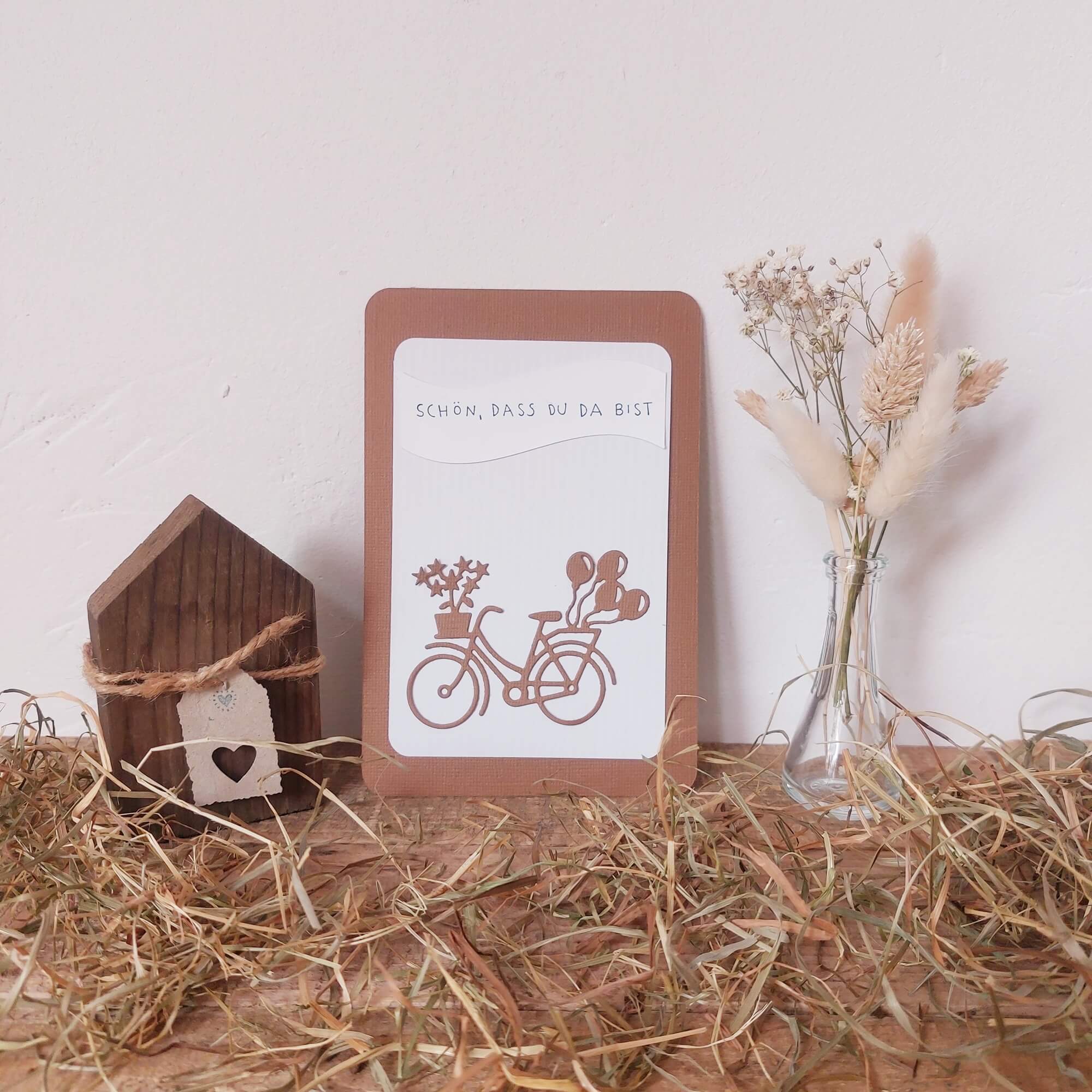 Eine handgefertigte Karte mit der Stanzenshop.de "Stanzschablone Fahrrad mit Blumen", gepaart mit einem Holzhaus und einer Trockenblumenvase auf Stroh, ergibt eine charmante Basteldeko - die perfekte Inspiration für Scrapbooking-Fans.