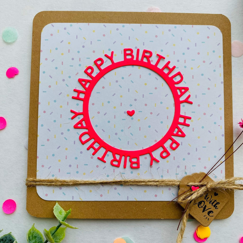 Eine Geburtstagskarte mit einer Stanzenschop.de-Stanzschablone „Happy Birthday auf rundem Schriftzug“ innerhalb eines roten kreisförmigen Rahmens, verziert mit buntem Konfetti und gebunden mit einer Bindfadenschleife mit einem hölzernen Herzanhänger.