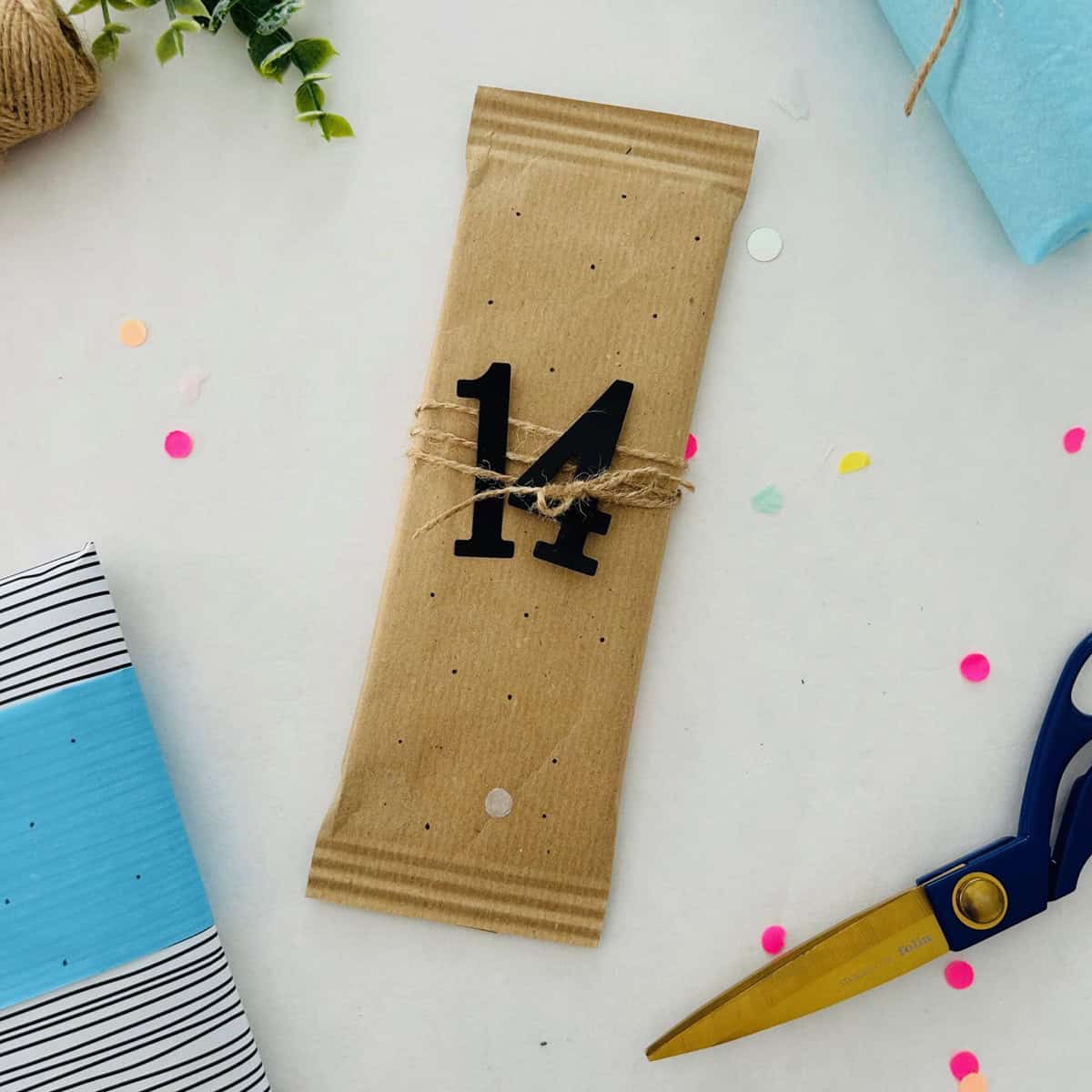 Ein rechteckiges, in braunes Papier eingewickeltes Geschenk mit der Zahl 14 aus dem Set Stanzschablone Große Zahlen von Stanzenshop.de, umgeben von Schere, Schnur, Konfetti und anderen Geschenken - ideal zum kreativen Basteln oder Scrapbooking.