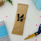 Ein rechteckiges, in braunes Papier eingewickeltes Geschenk mit der Zahl 14 aus dem Set Stanzschablone Große Zahlen von Stanzenshop.de, umgeben von Schere, Schnur, Konfetti und anderen Geschenken - ideal zum kreativen Basteln oder Scrapbooking.