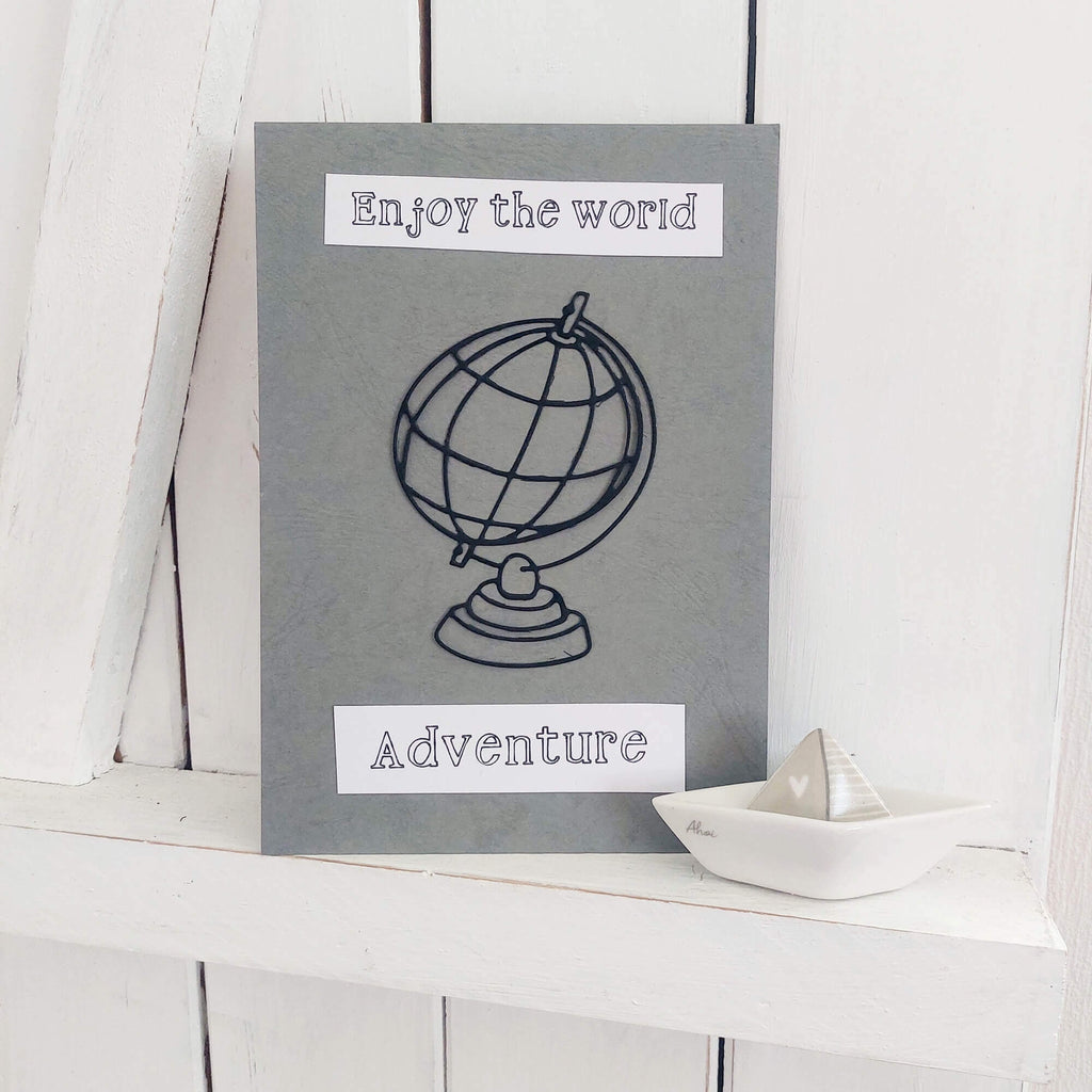 Eine Stanzenshop.de-Grußkarte mit den Worten „Die Welt genießen“ und „Abenteuer“ sowie die Illustration einer Stanzschablone: Großer Globus, platziert auf einer Holzoberfläche neben einem kleinen Keramikboot.