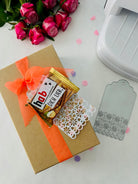 Eine Geschenkschachtel mit orangefarbenem Band, Pralinen, Danksagung, rosa Rosen, einer Stanzschablone Anhänger mit Verzierung von Stanzenshop.de (ideal für Scrapbooking) und einem dekorativen Anhänger steht auf einer weißen Fläche.