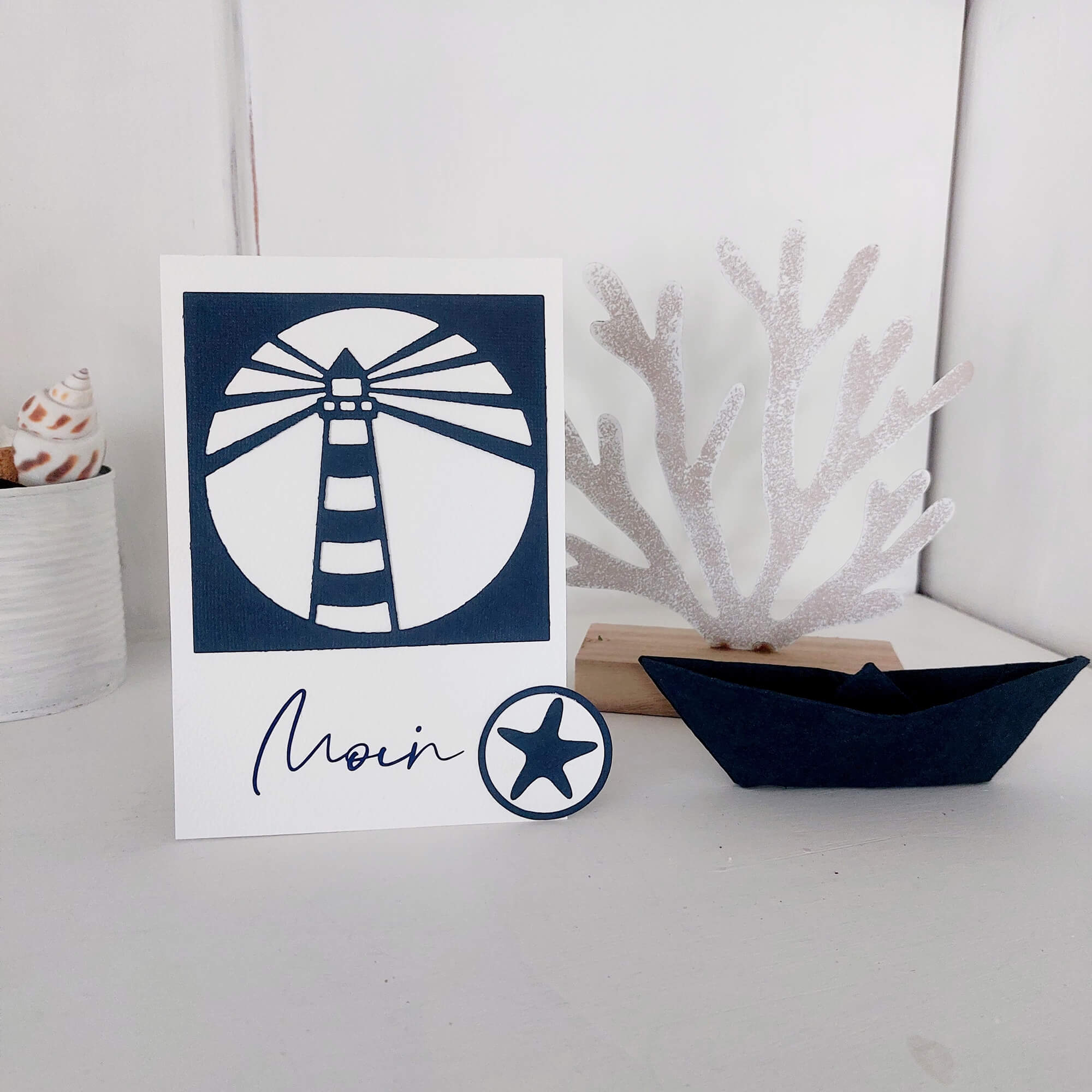 Eine Karte mit einem Leuchtturm aus der Stanzschablone Leuchtturm im Rahmen von Stanzenshop.de, mit "Moin", einer Seesterngrafik, einem schwarzen Papierboot, weißen Korallenakzenten und einer Muschel in einem weißen Topf auf weißem Hintergrund.