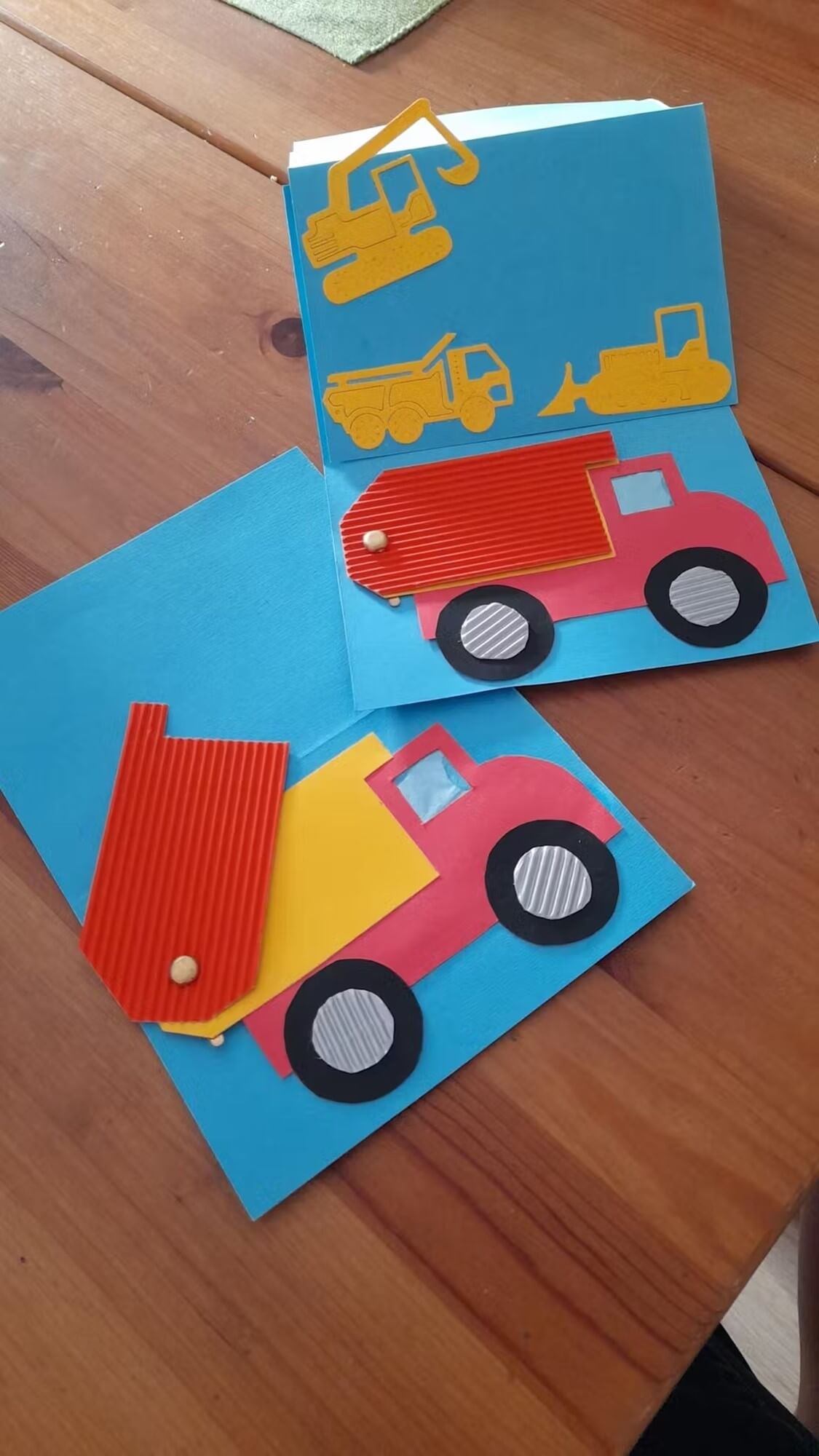Zwei handgefertigte Karten mit Stanzschablone Baufahrzeuge von Stanzenshop.de, mit Kippermotiven aus farbigem Papier und Wellpappe. Eine Karte ist offen und zeigt zusätzliche Fahrzeugformen auf einem Holztisch - ideal für Basteln-Fans.