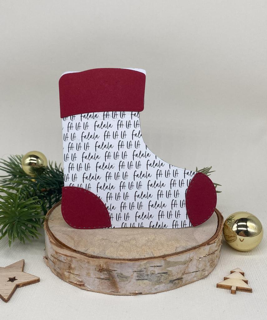 Eine Stanzschablone: Stiefelumschlag von Stanzenshop.de mit roten Akzenten und „fa la la“-Textmuster steht auf einer Holzscheibe, begleitet von Weihnachtsschmuck und Grün im Hintergrund. Perfekt für kreative Bastelprojekte, verleiht dieses charmante Stück jedem Raum eine festliche Note.