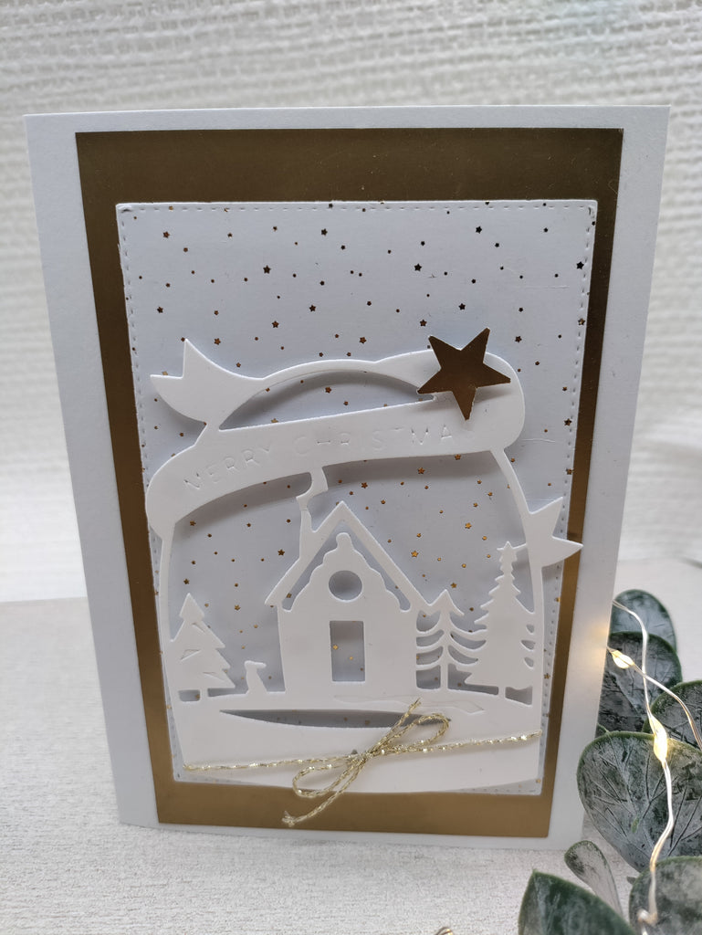 Eine handgefertigte Weihnachtskarte mit einer Stanzschablone: Haus in Schneekugel von Stanzenshop.de, mit weißen Ausschnitten eines Hauses, Bäumen und eines Sterns auf einem gold- und weißgesprenkelten Hintergrund, mit der Aufschrift „Frohe Weihnachten“ auf einem Banner. Perfekt für festliche Bastelprojekte oder als elegante Weihnachtsdekoration.