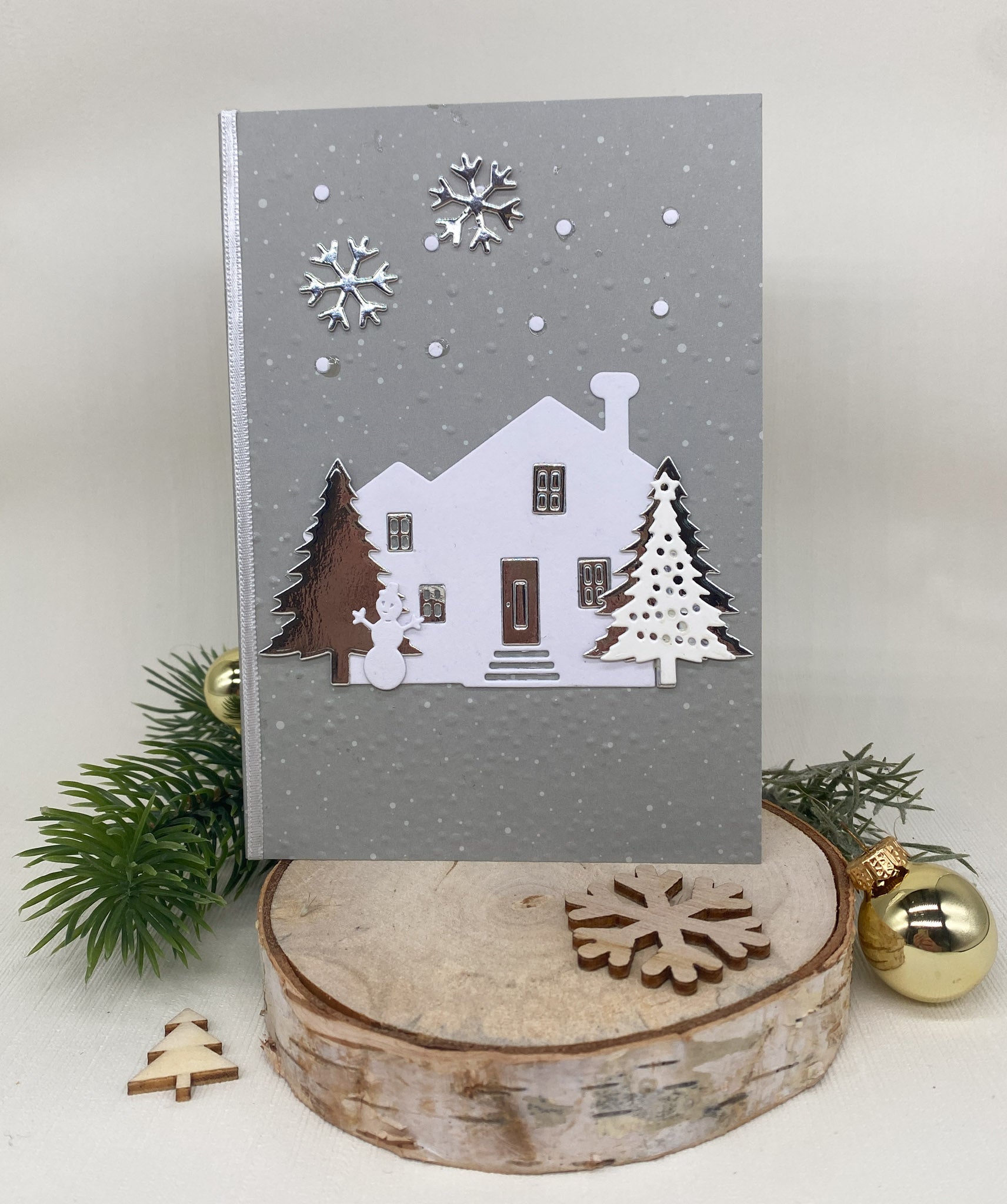 Eine graue Weihnachtskarte mit Stanzschablone: Haus mit Tannen von Stanzenshop.de, mit einem weißen Haus, Bäumen und einem Schneemann inmitten von Schneeflocken auf einem Holzständer mit Tannenzweigen - ideal für Scrapbooking oder Bastelprojekte.
