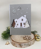 Eine graue Weihnachtskarte mit Stanzschablone: Haus mit Tannen von Stanzenshop.de, mit einem weißen Haus, Bäumen und einem Schneemann inmitten von Schneeflocken auf einem Holzständer mit Tannenzweigen - ideal für Scrapbooking oder Bastelprojekte.