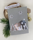 Eine graue Weihnachtskarte mit der Stanzenshop.de Stanzschablone: Haus mit Tannen, mit weißen Bäumen und Schneeflocken, auf einer Holzplatte mit Tannenzweigen, Ornamenten und einem "Schöne Weihnachten" Tag - ideal für Scrapbooking oder Bastelprojekte.