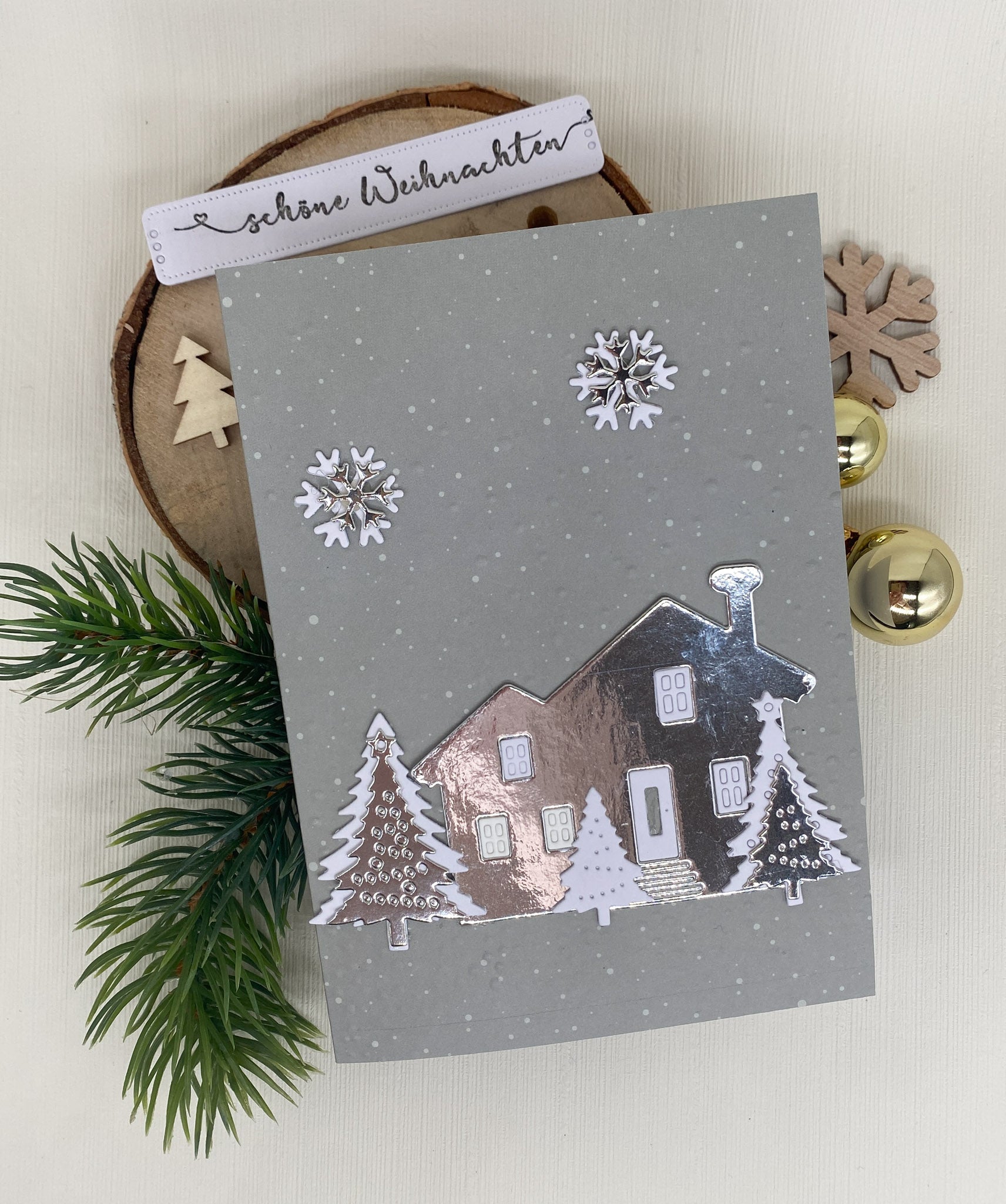 Eine graue Weihnachtskarte mit der Stanzenshop.de Stanzschablone: Haus mit Tannen, mit weißen Bäumen und Schneeflocken, auf einer Holzplatte mit Tannenzweigen, Ornamenten und einem "Schöne Weihnachten" Tag - ideal für Scrapbooking oder Bastelprojekte.