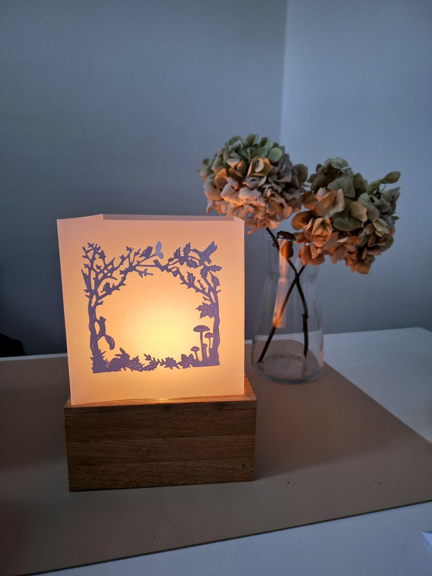 Eine Papierlaterne mit Herbstmotiven leuchtet auf einem Holzsockel neben getrockneten Hortensien und bietet die perfekte Inspiration für Bastelprojekte mit der Stanzschablone: Herbstwald von Stanzenshop.de auf einem weißen Tisch.