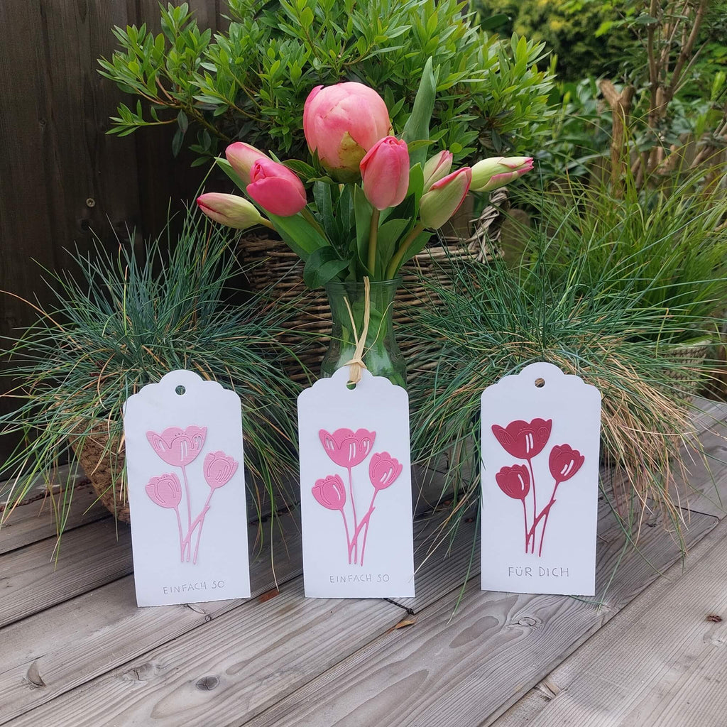 Drei Stanzschablone Drei Tulpen-Geschenkanhänger mit Tulpenmotiven und deutschem Text vor rosa Tulpen und grünem Blattwerk. Die Stanzschablone Drei Tulpen verleiht dem Design eine einzigartige Note.