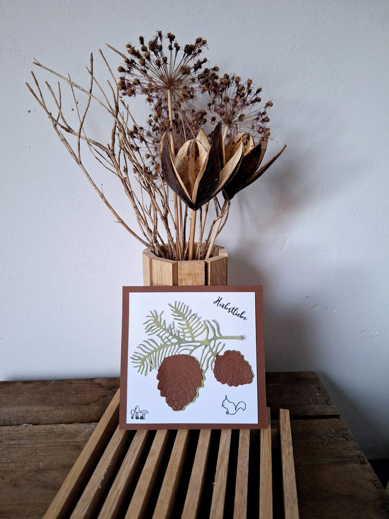 Getrocknetes botanisches Arrangement auf einem Holzblock gepaart mit einer illustrierten Karte auf einer Holzlattenoberfläche mit einer Stanzschablone: Zapfenzweig von Stanzenshop.de.