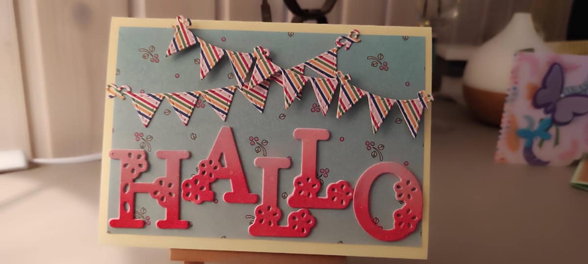 Eine handgefertigte Karte mit "HALLO" in fetten roten Buchstaben, erstellt mit der Stanzschablone: Blumenalphabet von Stanzenshop.de, zwei bunte Wimpelreihen und ein hellblauer Hintergrund, der mit kleinen Blumenmotiven verziert ist.