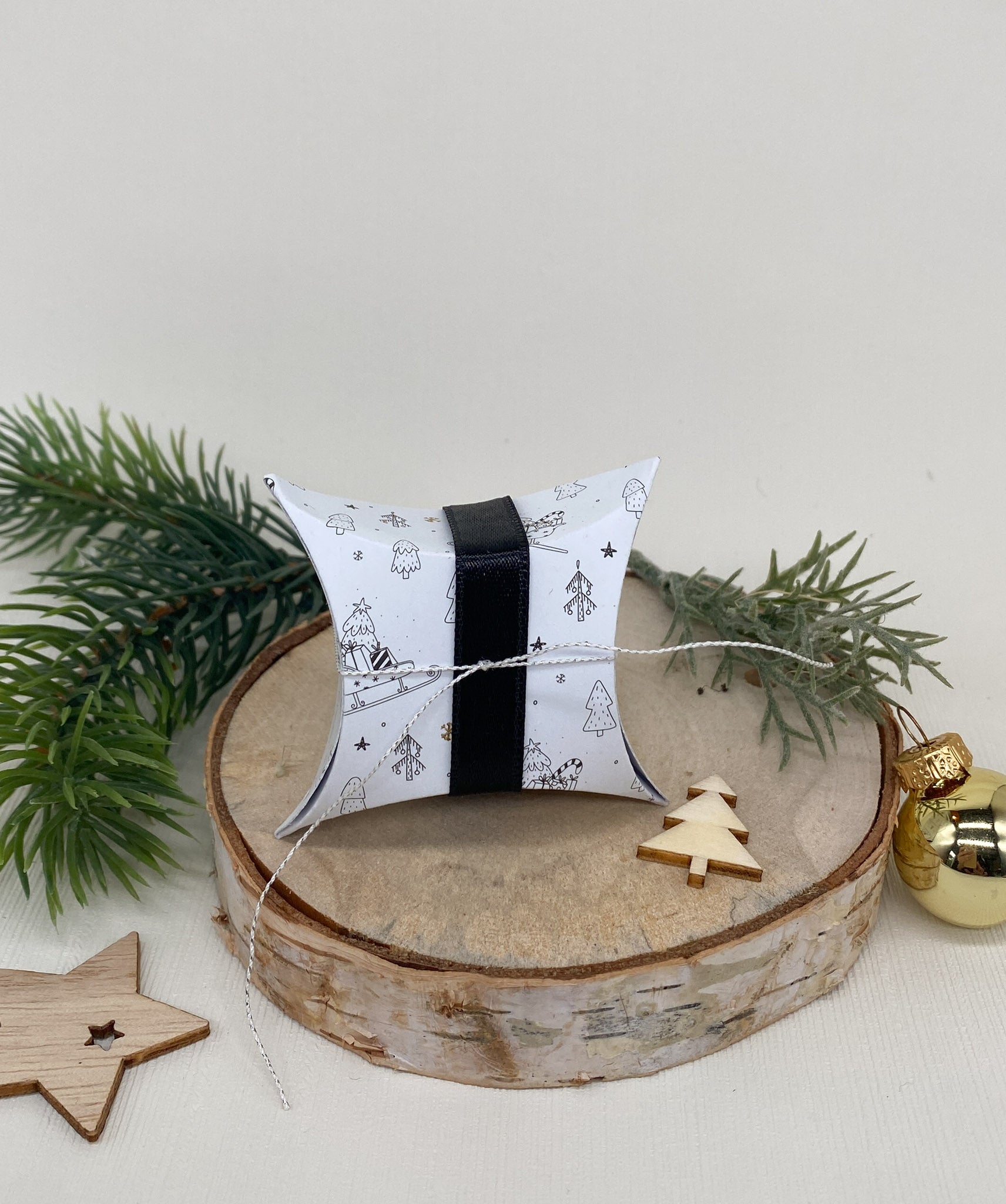 Eine Stanzschablone: Kissenschachtel von Stanzenshop.de mit schwarz-weißen Weihnachtsmotiven und einer schwarzen Schleife ruht auf Holz, umgeben von Tannenzweigen, Holzdekor und einem goldenen Ornament.