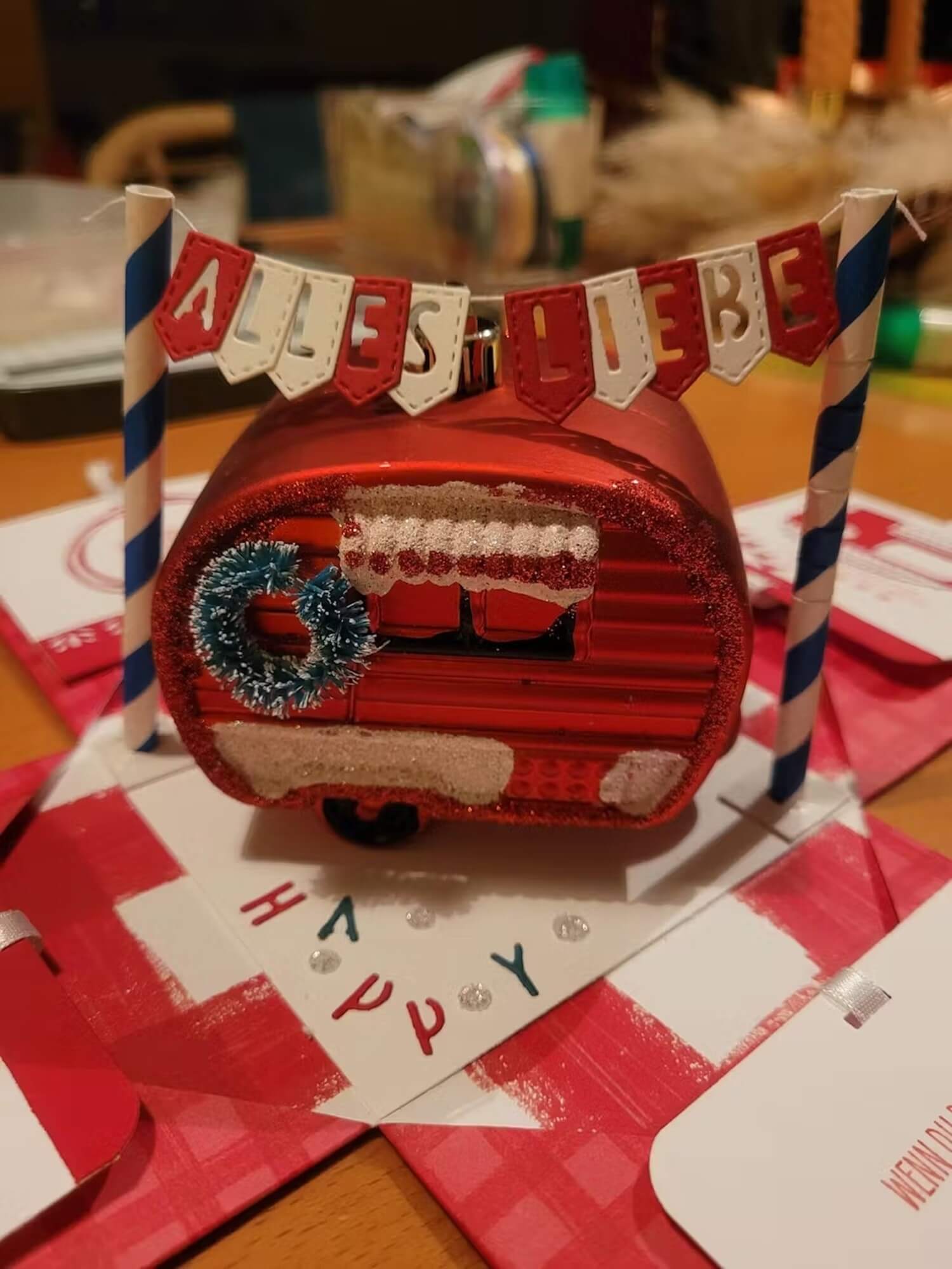 Ein rotes Ornament in Form einer Karawane mit Kranz und Schnee sitzt unter einem "ALLES LIEBE"-Banner. Für die Szene wurde die "Stanzschablone" von Stanzenshop.de verwendet: Wimpelkette mit Buchstaben", ideal für persönliche Kartendekorationen und Botschaften.