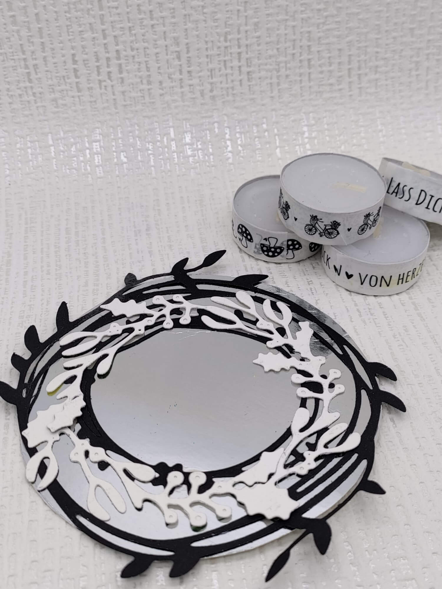 Die Stanzschablone Zwei Kreise mit Blumen von Stanzenshop.de ruht neben drei weißen Teelichtkerzen mit schwarzem Druckmotiv - ideale Akzente für Scrapbooking oder Kartenherstellung - auf einer strukturierten weißen Oberfläche.