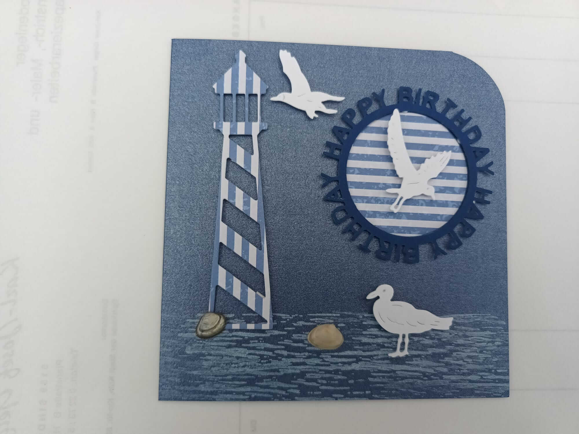 Eine blaue Geburtstagskarte mit einem gestreiften Leuchtturm, Kieselsteinen und Möwen, inspiriert von der Stanzschablone Drei Möwen von Stanzenshop.de, mit "HAPPY BIRTHDAY" in erhabenen Buchstaben um einen kreisförmigen Ausschnitt - perfekt für Scrapbooking.