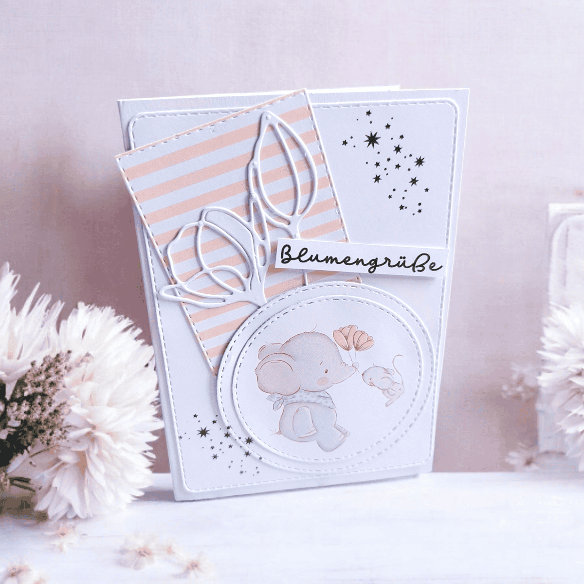 Eine Grußkarte mit einem Elefanten und einer Maus, floralen Akzenten, gestreiftem Hintergrund und "Blumengrüße", umgeben von weißen Blumen. Ideal zum Basteln von Karten mit der Stanzschablone Blume mit zwei Blüten von Stanzenshop.de; kompatibel mit Big Shot.