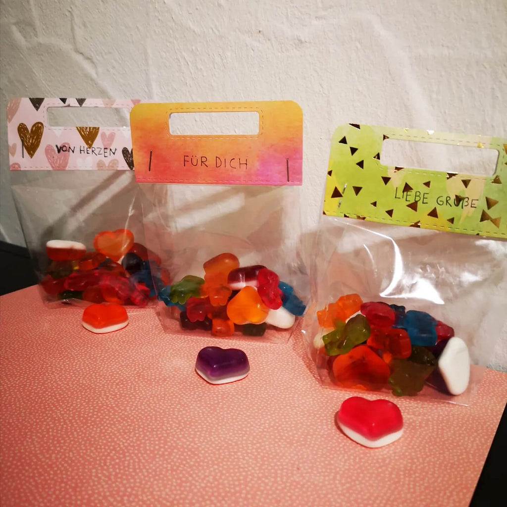 Drei kleine Plastiktüten gefüllt mit bunten Gummibonbons, auf denen jeweils die deutschen Aufschriften „Von Herzen“, „Für Dich“ und „Liebe Grüße“ stehen. Diese Bonbons sind perfekt für ein Scrapbooking-Projekt und verleihen dem Ganzen eine süße Note. Einige Bonbons sind vorne verstreut und bereit, mit Ihrer Stanzschablone Griffe von Stanzenshop.de kombiniert zu werden.