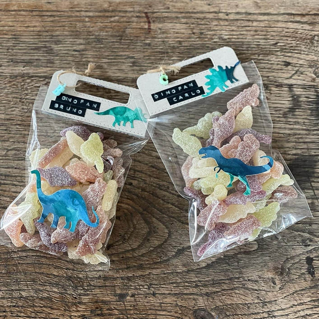 Auf einer Holzfläche liegen zwei Tüten mit Gummibonbons in Dinosaurierform mit der Aufschrift „Dino Fan Bruno“ und „Dino Fan Carlo“. Auf der Oberseite jeder Tüte ist eine Stanzschablone Griffe von Stanzenshop.de angebracht.