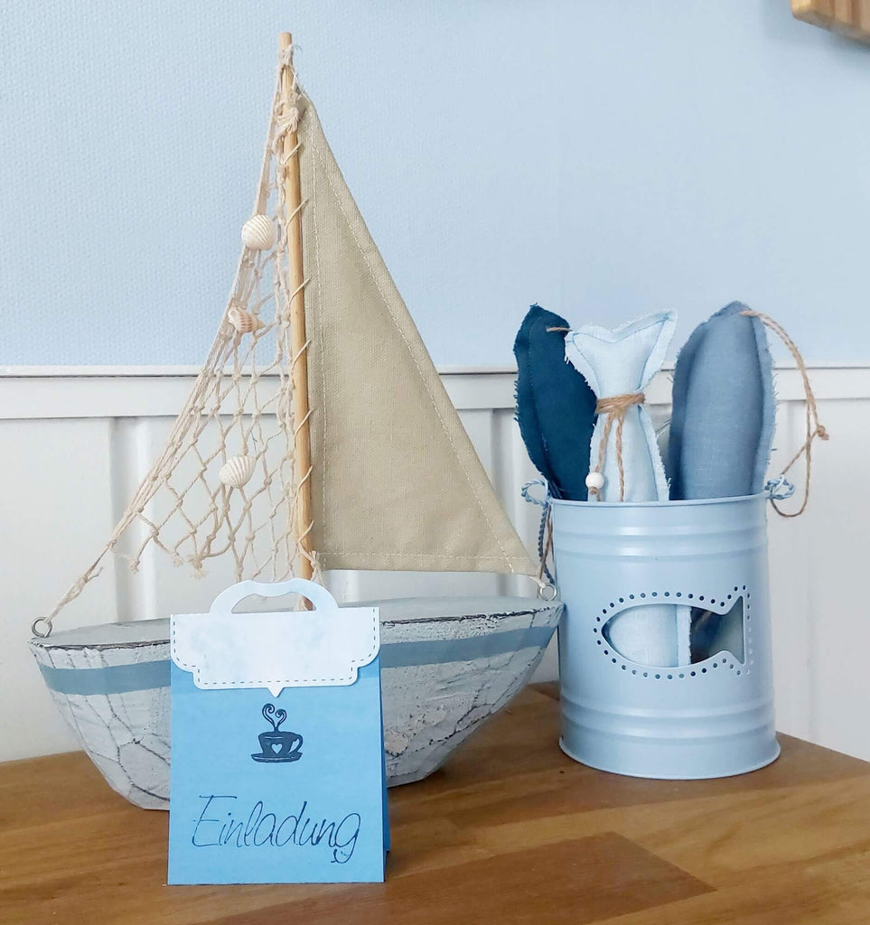 Auf einem hellblau-weißen Untergrund stehen auf einer Holzfläche ein dekoratives Segelboot, ein blauer Eimer mit Stofffischen und eine Einladungskarte mit der Aufschrift „Einladung“, gebastelt mit der Stanzschablone Verschluss für Tüten von Stanzenshop.de.