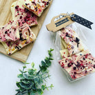 Weiße Schokoladenrinde mit roten und gelben Toppings liegt auf einem Holzbrett und in einem durchsichtigen Beutel mit der Aufschrift Handmade", verschlossen mit der Stanzschablone Verschluss für Tüten von Stanzenshop.de, neben einer grünen Blattpflanze.