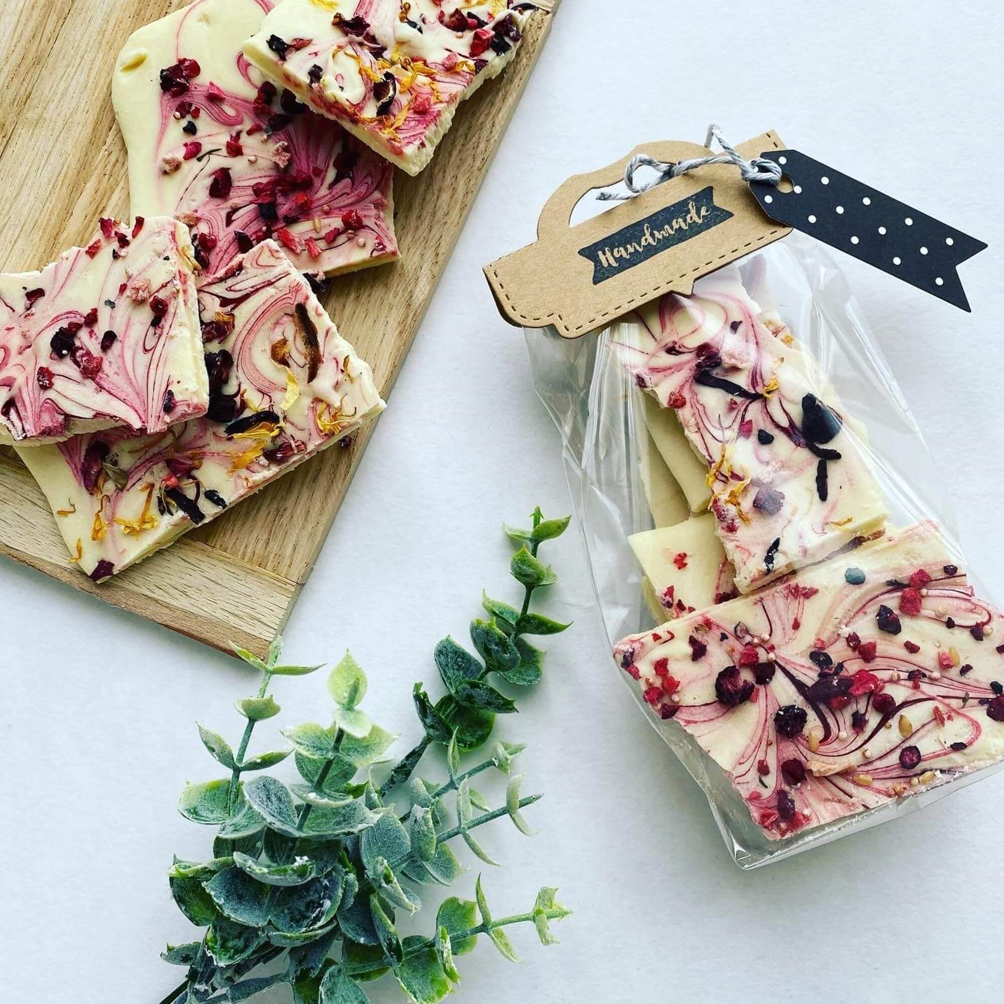 Weiße Schokoladenrinde mit roten und gelben Toppings liegt auf einem Holzbrett und in einem durchsichtigen Beutel mit der Aufschrift Handmade", verschlossen mit der Stanzschablone Verschluss für Tüten von Stanzenshop.de, neben einer grünen Blattpflanze.
