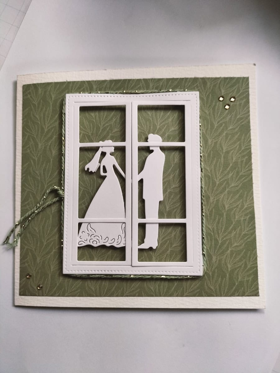 Eine Hochzeitskarte mit weißer Silhouette von Braut und Bräutigam, gefertigt mit der Stanzenshop.de Stanzschablone Brautpaar und umrahmt von einem Papierfenster auf grünem strukturierten Hintergrund - ideal für Basteln oder stilvolle Hochzeitseinladungen.
