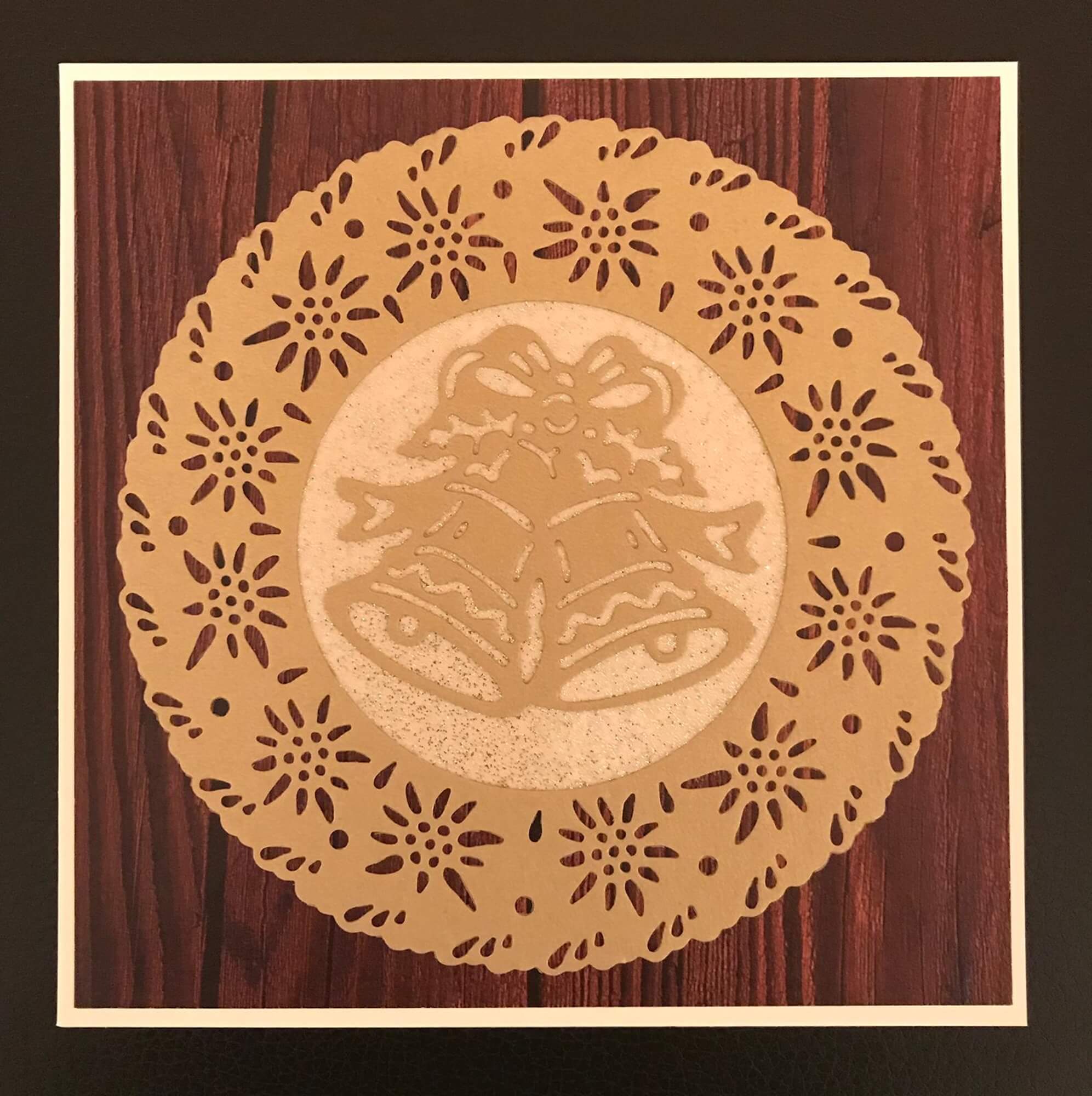 Ein rundes Papierdeckchen mit einem Ausschnitt von zwei Glocken und einer Schleife, hergestellt mit der Stanzenshop.de Stanzschablone: Zwei Weihnachtsglocken, ausgestellt auf einer Holzfläche.