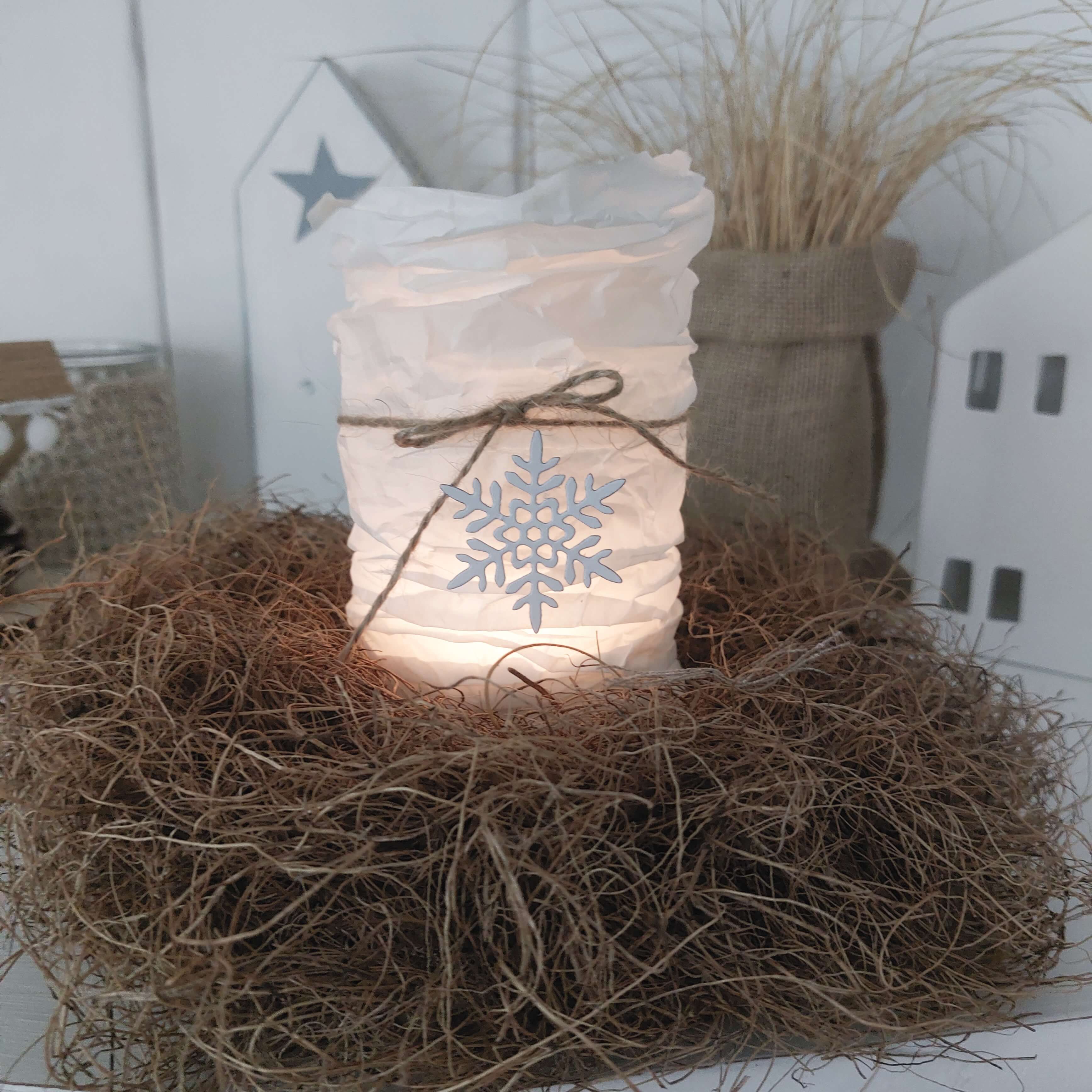Eine brennende Kerze in einem weißen, zerknitterten Halter mit einer Schleife aus Garn und dem Dekor "Stanzschablone Vier Schneeflocken in verschiedenen Größen" von Stanzenshop.de, perfekt zum Basteln oder Scrapbooking, platziert in getrocknetem Gras.