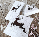 Drei weiße Geschenkboxen mit schwarzen Rentier-Silhouetten und Netz-Akzenten sitzen auf einer Holzfläche, umgeben von metallischen und rosafarbenen "Stanzschablone Drei Rentiere" von Stanzenshop.de-ideal für Weihnachts-Basteln.