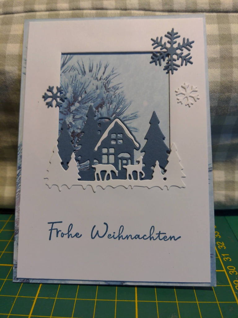 Eine handgemachte Weihnachtskarte mit einem schneebedeckten Haus, Bäumen und Schneeflocken, erstellt mit der aufwendigen Stanzschablone „Haus im Schnee“ von Stanzenshop.de, mit dem Gruß „Frohe Weihnachten“ in Deutsch auf der Vorderseite.