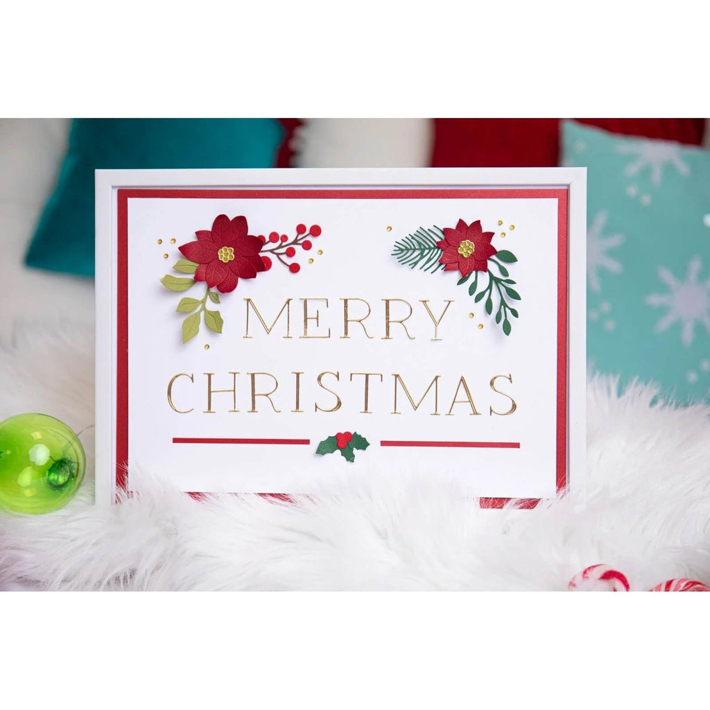 Eine „Frohe Weihnachten“-Karte mit roten Blumen und grünen Blättern ist auf weichem weißem Stoff abgebildet, akzentuiert durch Weihnachtsdekorationen im Hintergrund. Perfekt für Scrapbooking-Fans, die das Sizzix Thinlits Die Set Festive Foliage verwenden, verleiht diese Karte Ihren Weihnachtsprojekten festlichen Charme.