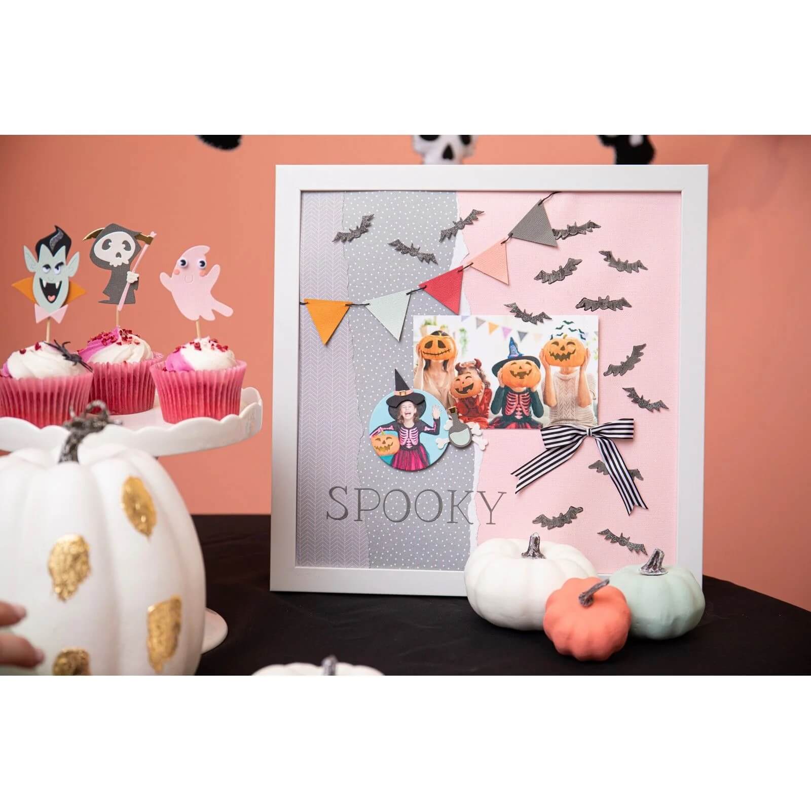 Eine gerahmte Halloween-Collage zeigt die Sizzix 666453 - Thinlits Spooky Icons von Sizzix, zusammen mit Cupcakes, weißen und pastellfarbenen Kürbissen und Fledermausausausschnitten auf einem schwarzen Tisch vor einem pfirsichfarbenen Hintergrund.