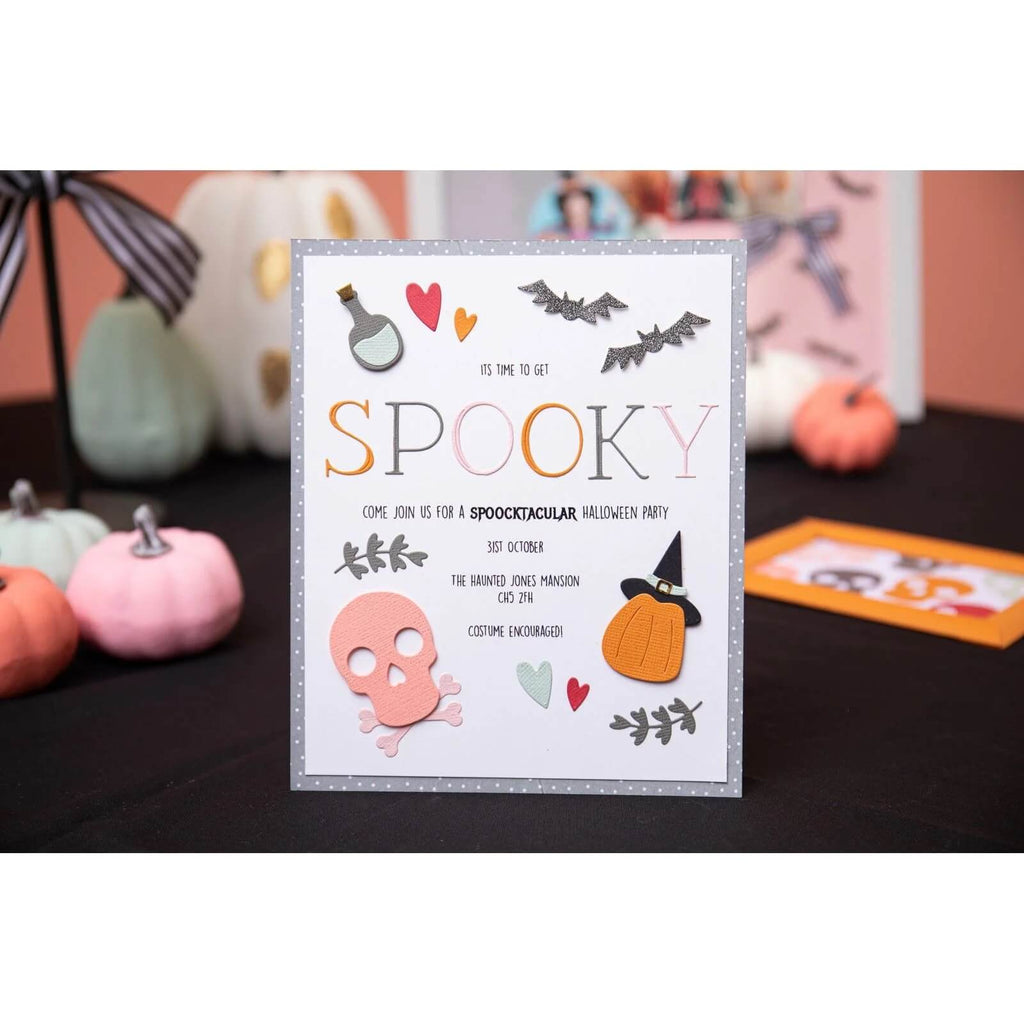 Eine Einladungskarte zu einer Halloween-Party mit Text, Fledermausdekorationen, einem rosa Totenkopf, der mit dem Sizzix Thinlits Die Set Spooky Icons von Sizzix erstellt wurde, einem Kürbis und Herzillustrationen, angezeigt mit Kürbissen im Hintergrund.